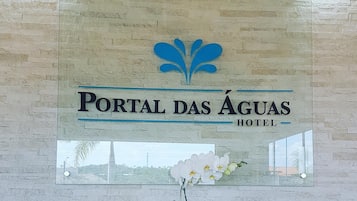Placa do Hotel Portal das Águas com logotipo, uma orquídea em vaso e uma janela refletindo uma paisagem.