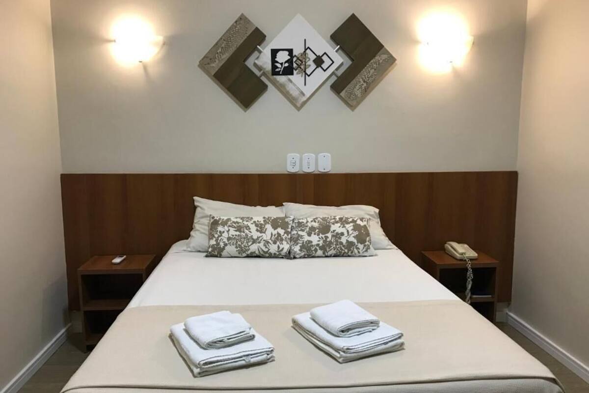 Een netjes opgemaakt bed met bloemenkussens, twee nachtkastjes met lampen, en een wandlamp.