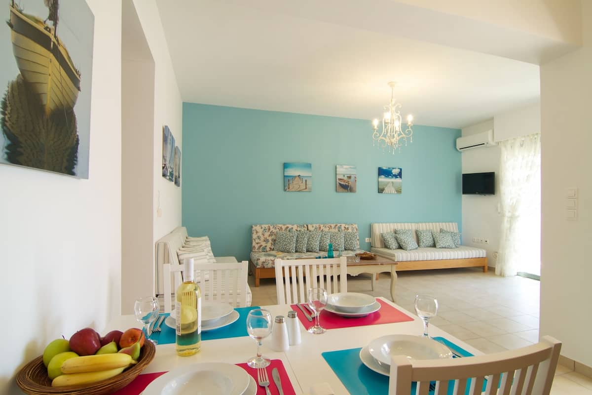 A dining area with a table set for two, a fruit bowl, and a sofa in the background.
