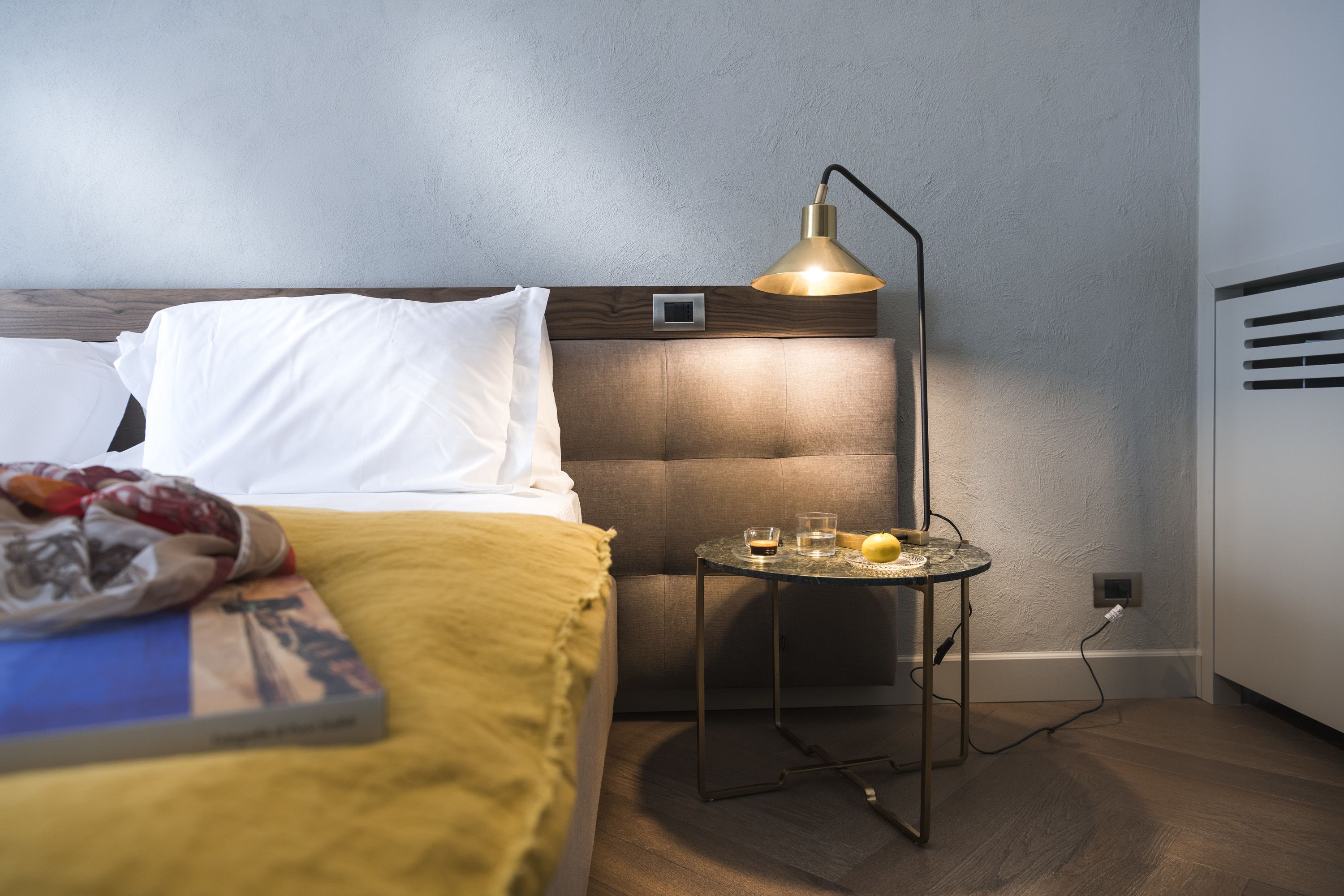 Un lit bien fait, avec des oreillers blancs et une couverture jaune, une table de chevet avec une lampe, et une petite table sur laquelle se trouvent un verre et un fruit.