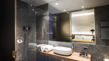 A modern bathroom with a glass-enclosed shower, a wooden vanity with a sink, and a large mirror reflecting a bed and a tripod.