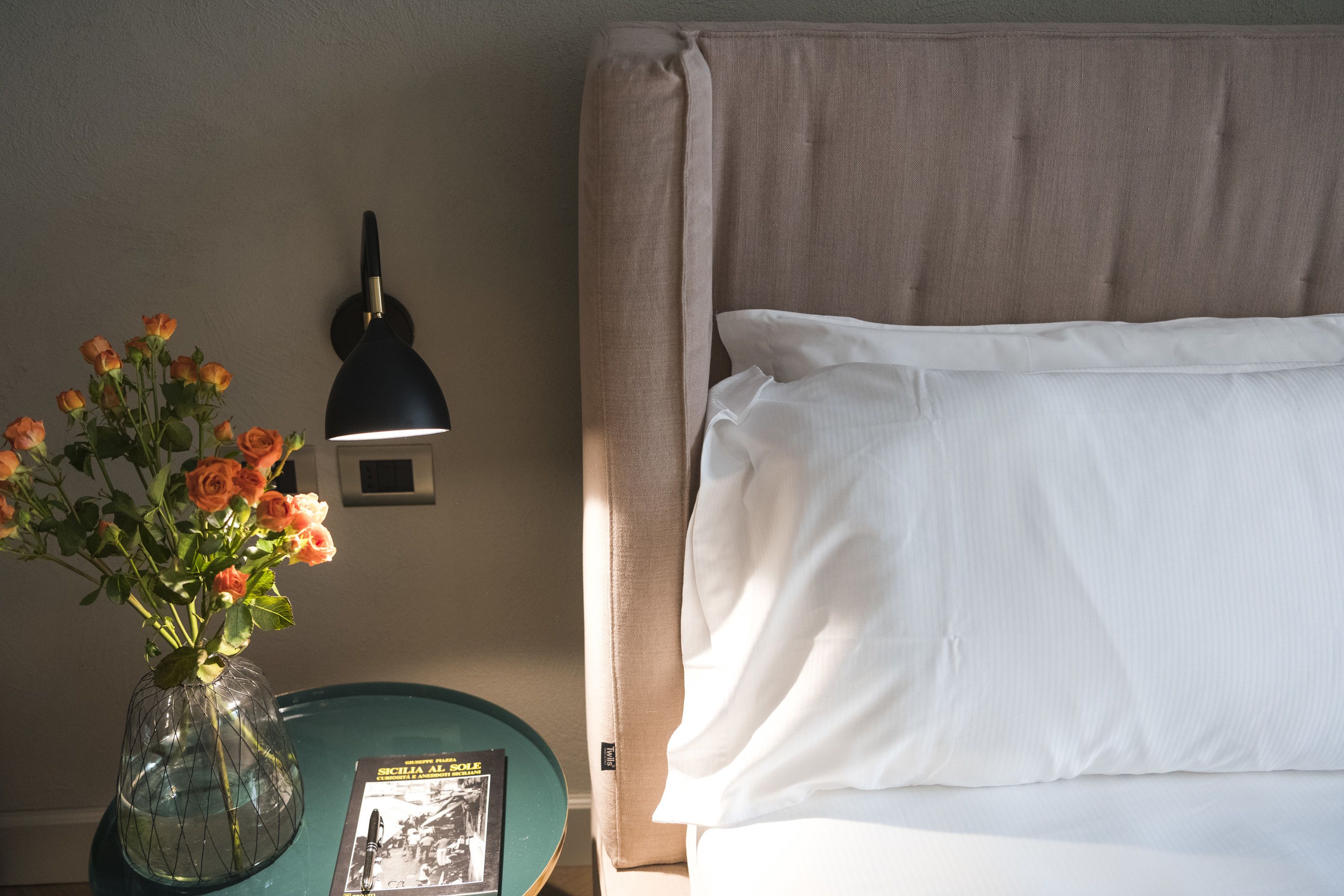A neatly made bed with white linens, a vase with orange roses, a black desk lamp, and a small book on a teal side table.