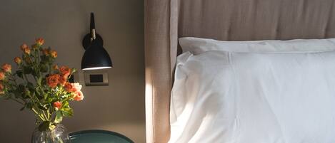 A neatly made bed with white linens, a vase with orange roses, a black desk lamp, and a small book on a teal side table.