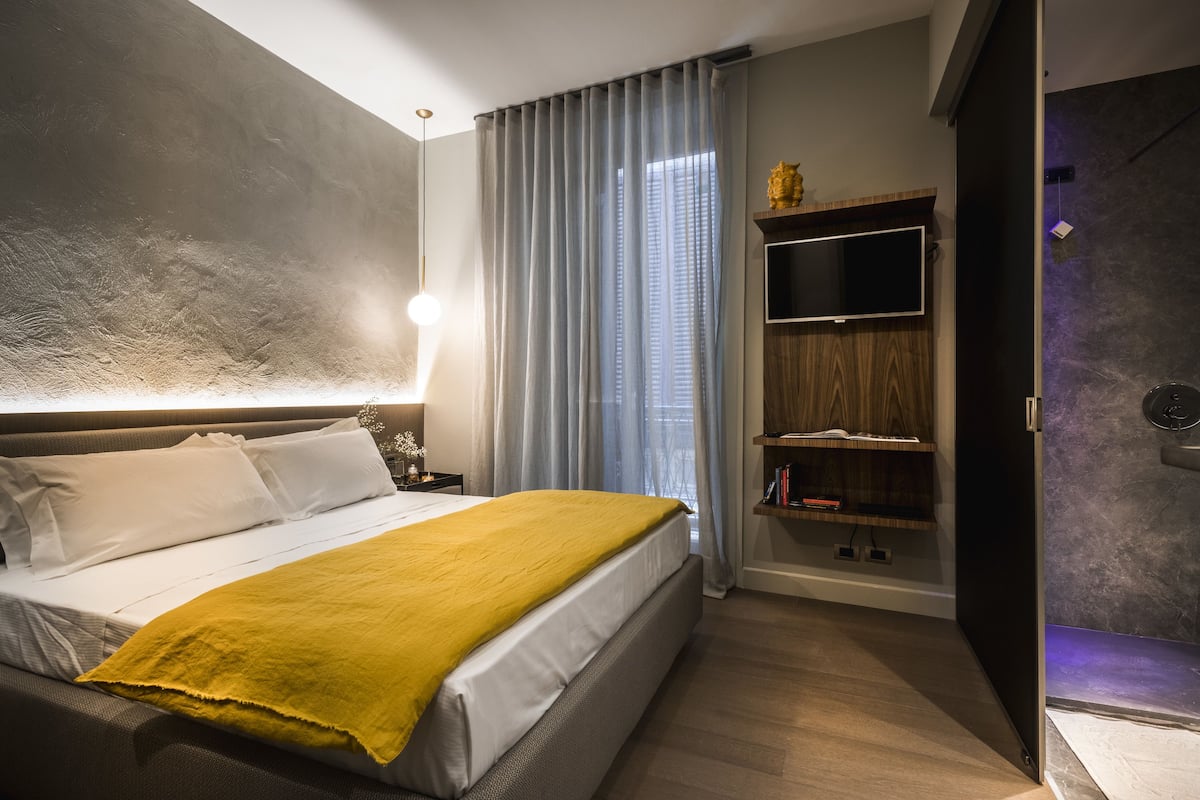 Une chambre à coucher moderne avec un grand lit, un téléviseur fixé au mur et une étagère en bois remplie de livres.