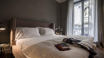 Una moderna camera d'albergo con un letto grande, un comodino con una lampada, una finestra che si affaccia su una vista cittadina e un tavolino con un libro e dei bicchieri.