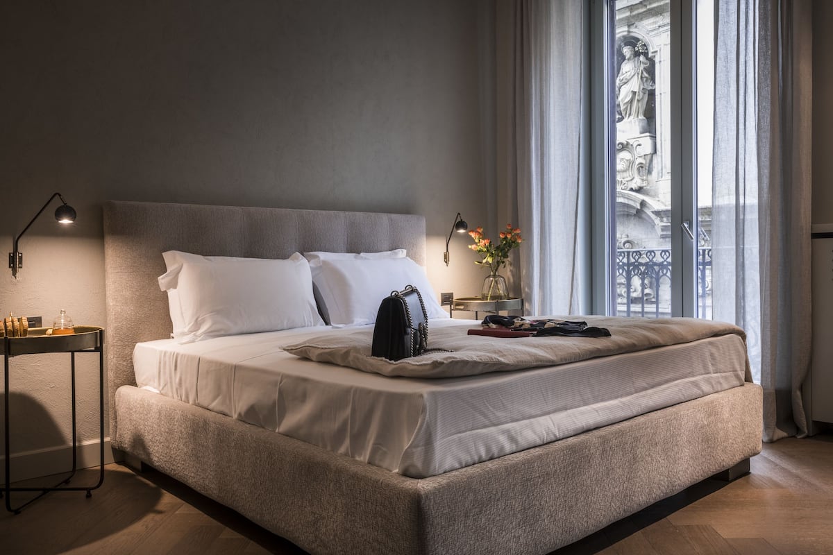 Un lit bien fait, avec du linge de lit blanc, une table de chevet avec une lampe, et une vue sur une statue par la fenêtre.