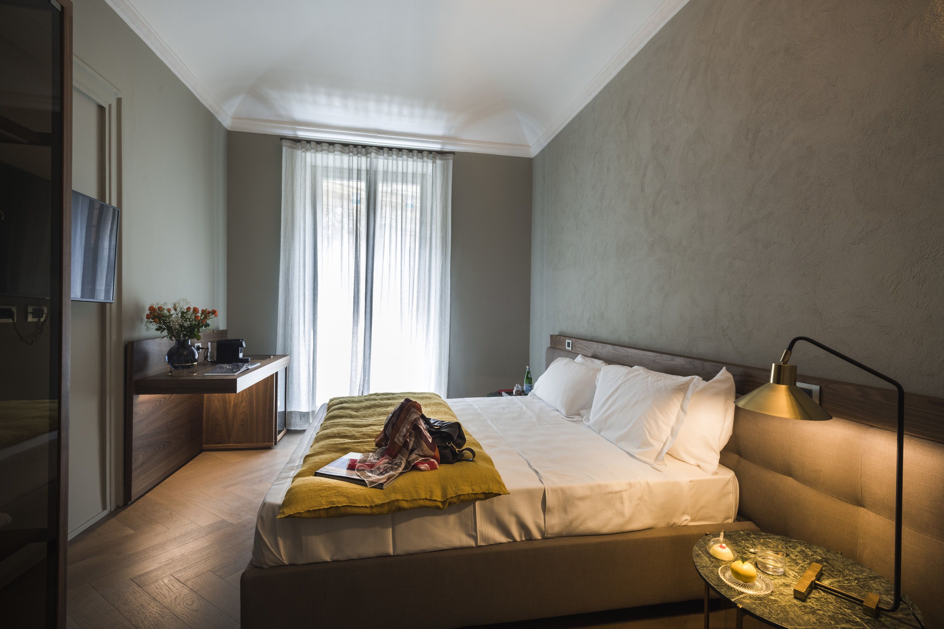 A hotel room with a bed, bedside table, lamp, and a view of the outdoors through the window.