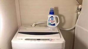 A compact, white, wall-mounted, single-occupant washing machine with a control panel and a detergent bottle on top.