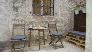 Un muro de piedra rústica con una ventana de metal, dos sillas de madera con cojines, una mesa pequeña con un plato de comida y tazas, y un banco de madera con un cojín.