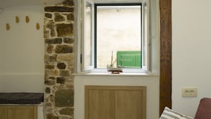 Una habitación con pared de piedra, una ventana con vista a un portón verde, un armario de madera y un cojín morado.
