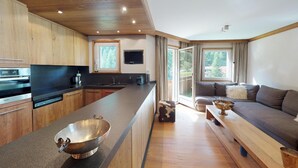 Een moderne keuken met houten kastjes, een inbouwoven en een groot eiland met een metalen kom. Er is een bank en een bijzettafeltje in een woonruimte met een groot raam.