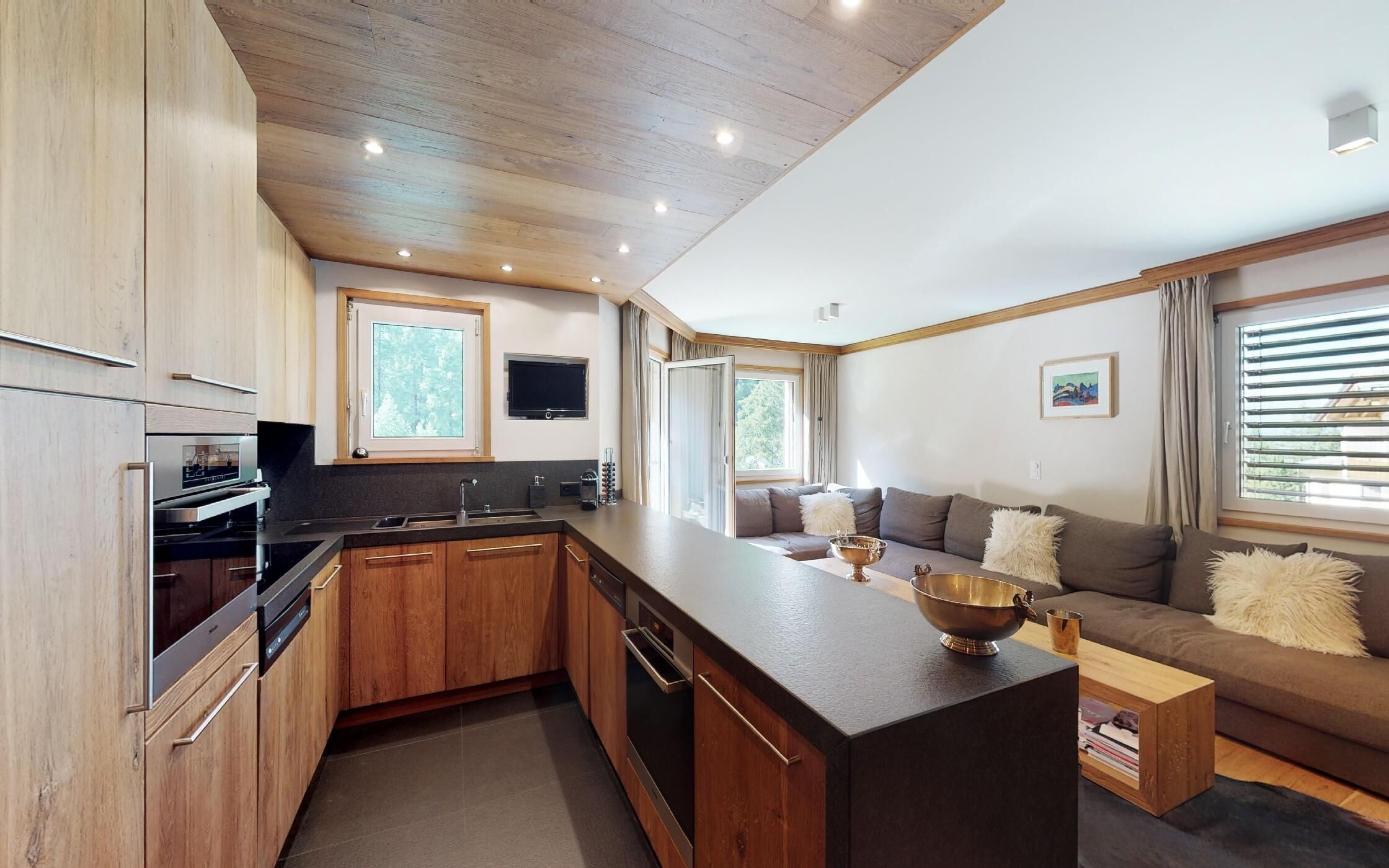 A modern kitchen with wooden cabinets, a built-in oven, and a large island. There is a seating area with a sofa and a coffee table.