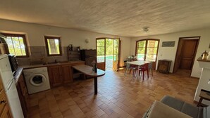 Una cocina con lavadora, armarios de madera, una mesa de comedor con sillas y vistas al exterior a través de ventanas amplias.