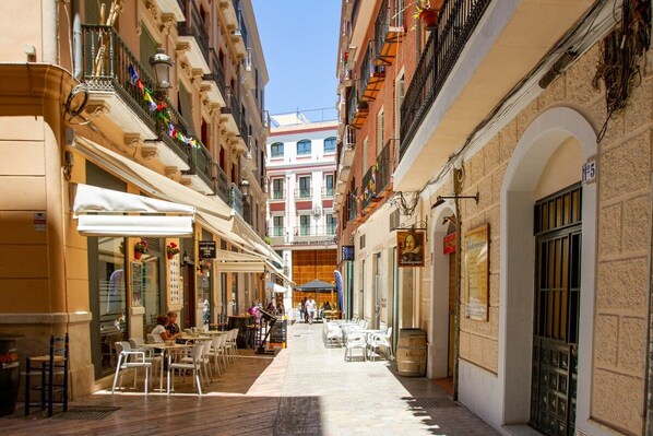 A narrow street with outdoor seating, buildings with balconies, and a clear sky.