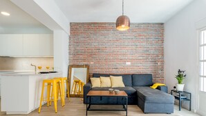 A modern living space with a kitchen, a grey sofa, yellow stools, and a brick wall. - Julia Centro Historico (Málaga)