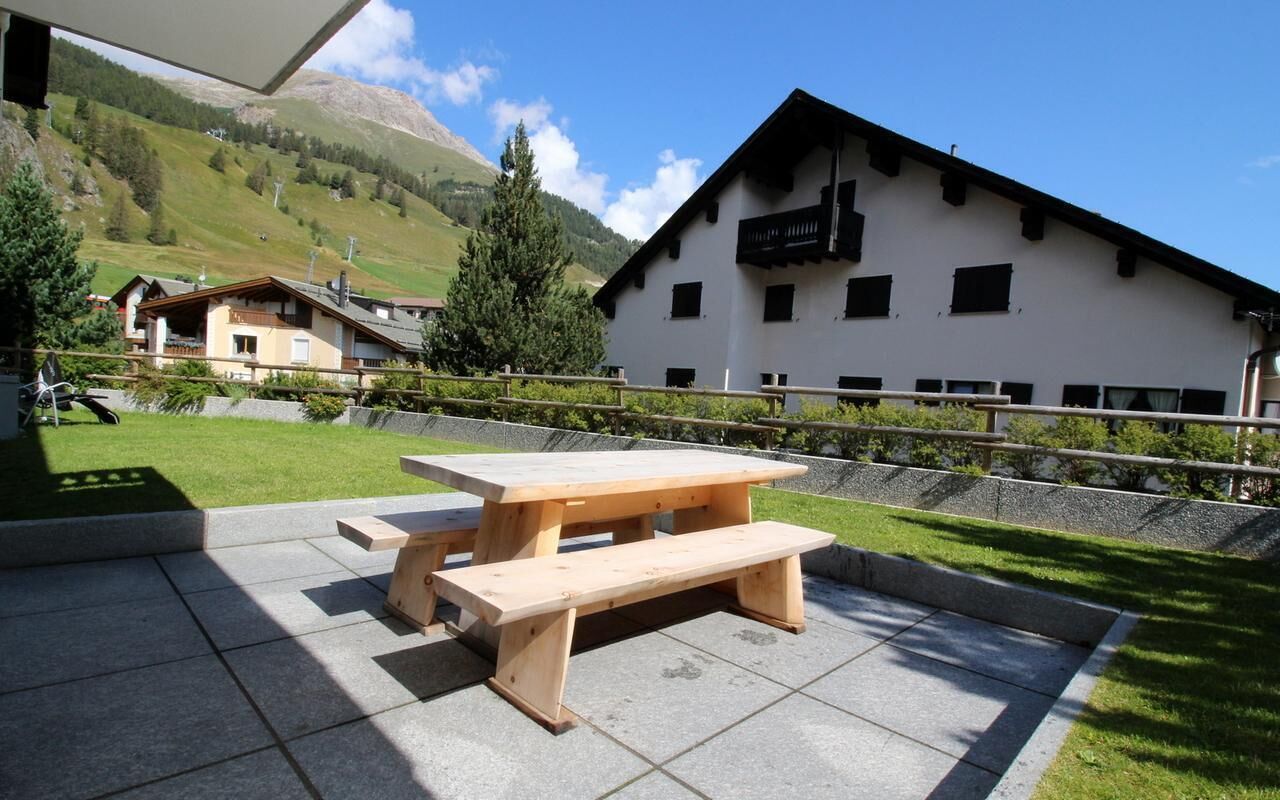 Une table de pique-nique en bois, avec des bancs, située dans une cour dont l’arrière-plan est constitué d’une chaîne de montagnes.