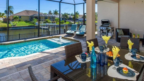 A poolside dining area with a view of a swimming pool and a house in the background. - Coral Paradise- Salt Water Pool - baby fence (Cape Coral)