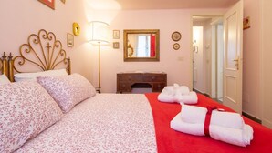 Un letto rifatto con lenzuola a motivi floreali, una lampada da comodino, una cassettiera in legno e una coperta rossa.