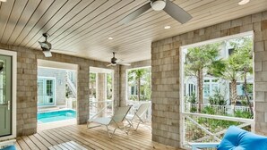 Un espacio al aire libre techado con suelo de madera, mobiliario blanco y vistas a una piscina y zonas verdes.