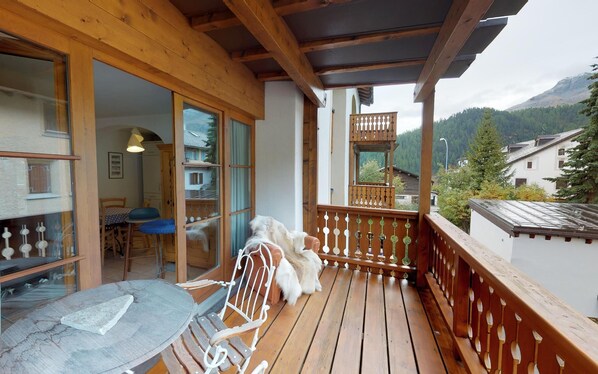 A wooden balcony with a table and chairs, a dog, and a view of a mountain and houses.