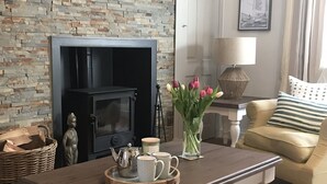 Un salon avec un mur en pierre, un poêle à bois noir, un canapé, une table basse avec un plateau, un vase de tulipes et une lampe.