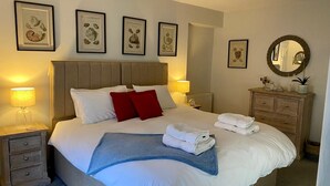 Un lit bien fait, avec du linge de lit blanc, des coussins rouges et un plaid bleu. Des tableaux encadrés ornent le mur, ainsi qu’une commode en bois et un miroir rond.