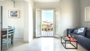 Uma sala de estar com sofá, uma mesa de centro com garrafa e copos, uma mesa de jantar com cadeiras, e vista para área externa através de uma porta aberta.