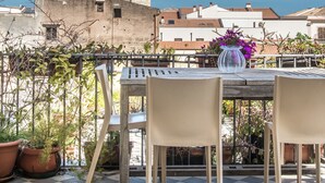 Um terraço com chão xadrez, duas cadeiras, uma mesa com um vaso de flores e vista para edifícios e uma torre ao fundo.