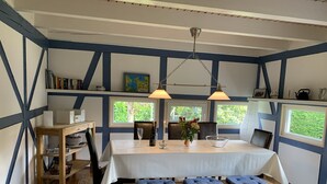 A dining area with a wooden table, benches, and a rug. There are bookshelves and a window with a view of greenery.