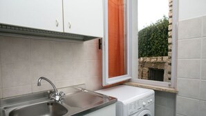 A compact kitchen with a stainless steel sink, a built-in washing machine, and a window with an orange curtain. - M084 - Marcelli, three-room apartment with garden at Residence MareVerde (Marcelli)