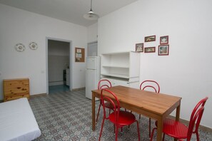A room with a bed, a wooden table with red chairs, a white refrigerator, and a door leading to another room.