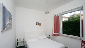 A single bed with white bedding, a small bedside table, a hanging light fixture, a window with a red curtain, and a framed picture on the wall. - M084 - Marcelli, three-room apartment with garden at Residence MareVerde (Marcelli)