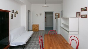 A small, clean room with a wooden table, a white sofa, and a red chair. There is a door leading to another room, a small wooden cabinet, and a white shelf unit. - M084 - Marcelli, three-room apartment with garden at Residence MareVerde (Marcelli)