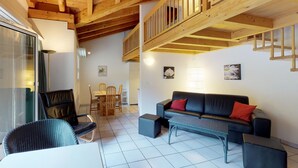Una sala acogedora con techo de madera, un sofá negro con cojines rojos, una mesa de comedor con sillas y un balcón con barandas de madera.