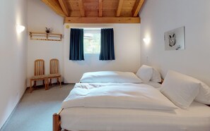 A bedroom with a wooden ceiling, two single beds, a window with curtains, a wall-mounted shelf with a plant, and a framed picture.