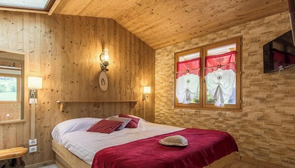 Een houten kamer met een bed, een wandlamp, een raam met gordijnen, en een televisie aan de muur.