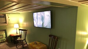 A room with a television mounted on the wall displaying an image, two wooden chairs, a small table with a lamp, and a carpeted floor. - Rustic Beach Front Apartment - Cozy, Rustic and On the Beach! - 2 BR/1 Bath (Norfolk)