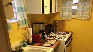 A compact kitchen with a gas stove, a toaster, and a microwave. There is a sink with a dish rack and a trash can. The kitchen has wooden cabinets and a window with curtains. - Rustic Beach Front Apartment - Cozy, Rustic and On the Beach! - 2 BR/1 Bath (Norfolk)