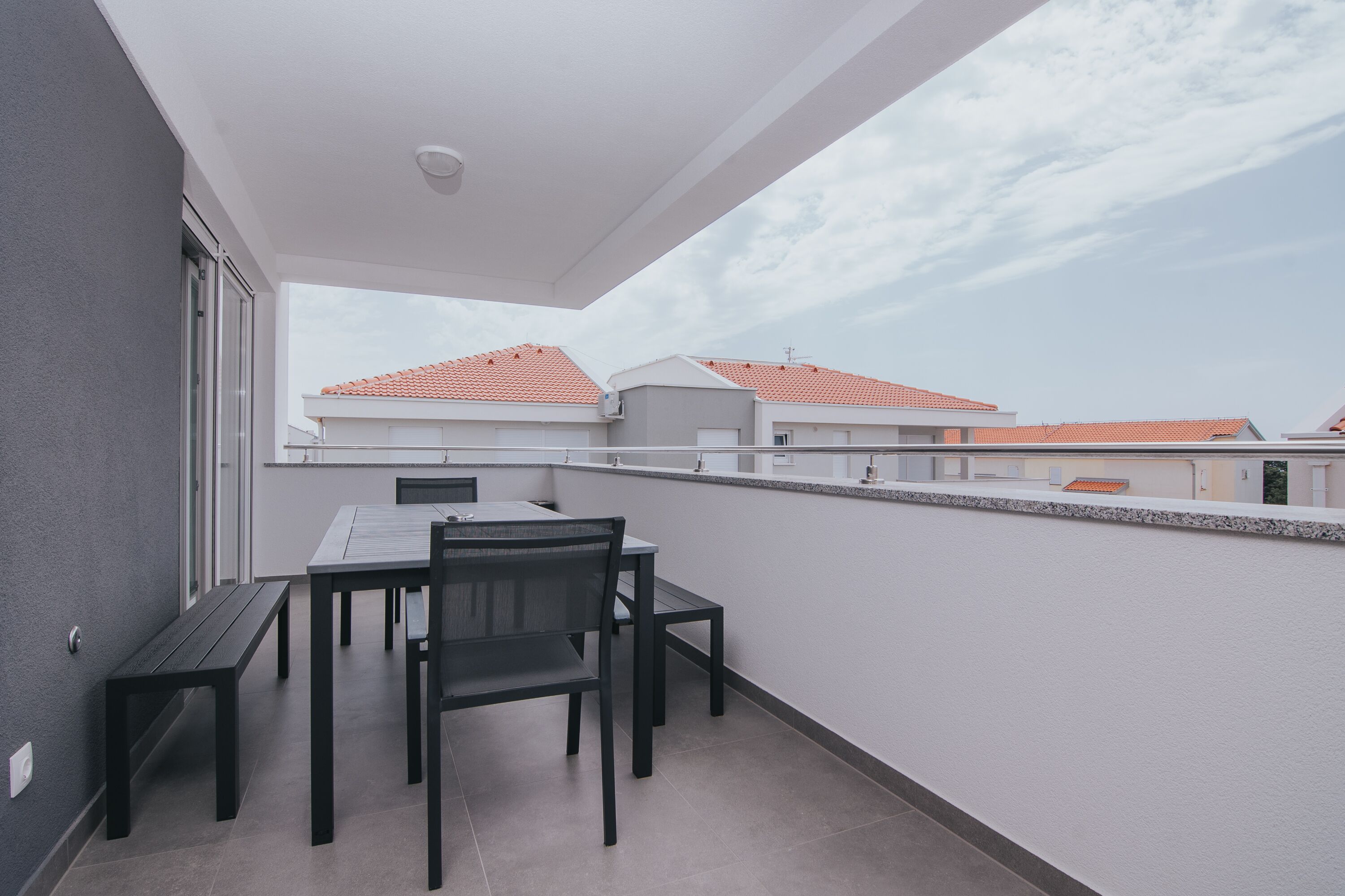 Un balcon avec une table et des chaises, donnant sur un quartier résidentiel aux toits recouverts de tuiles rouges.