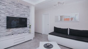 Ett modernt vardagsrum med en platt-TV som är monterad på en vägg med en intressant yta, en vit soffa med svarta kuddar och ett vitt soffbord med en dekorativ detalj.