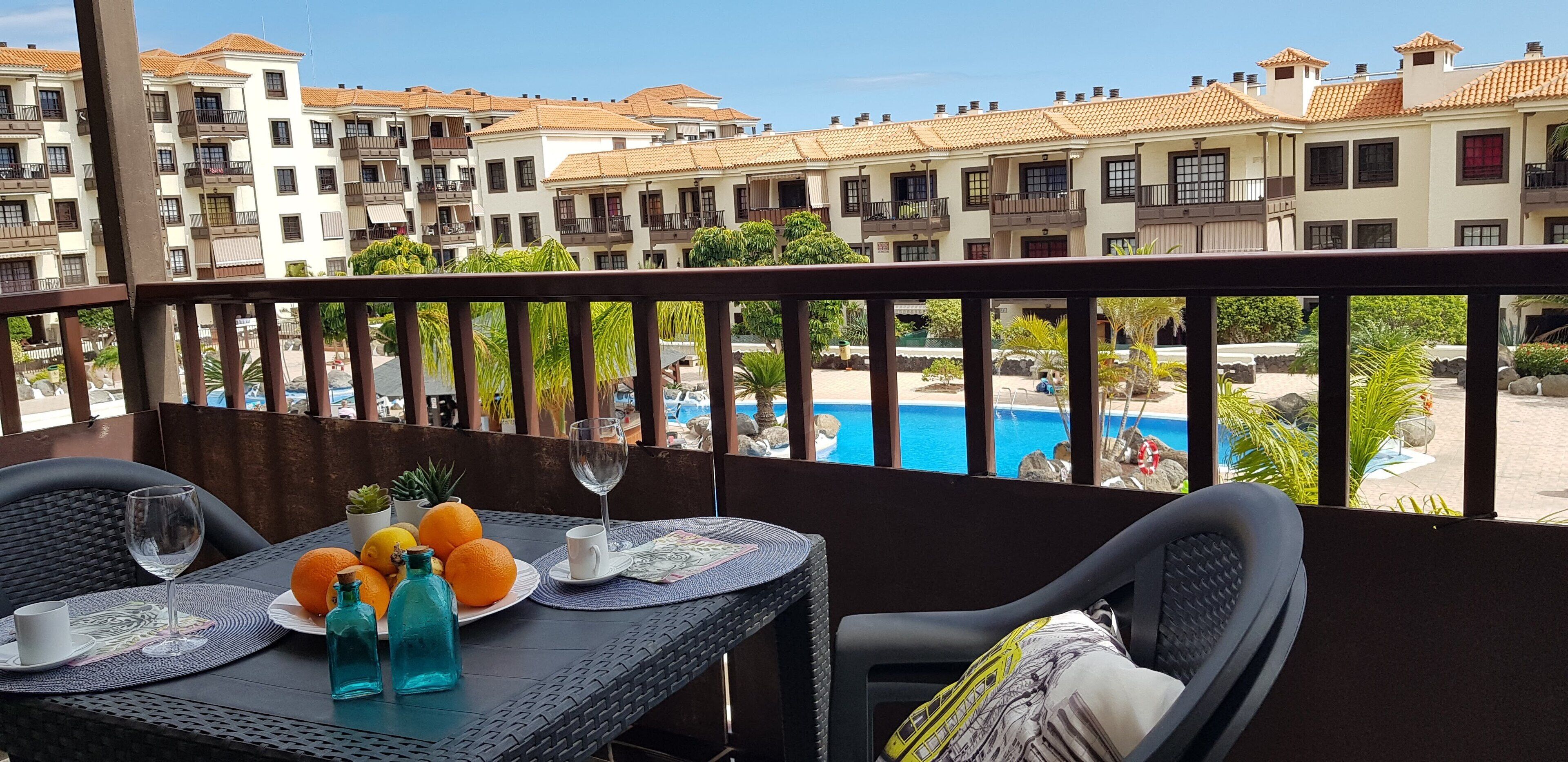 A balcony with a table set for two, overlooking a swimming pool and a building complex.