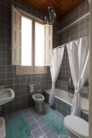 Et badeværelse med toilet, bidet, badekar og et vindue med gardiner.