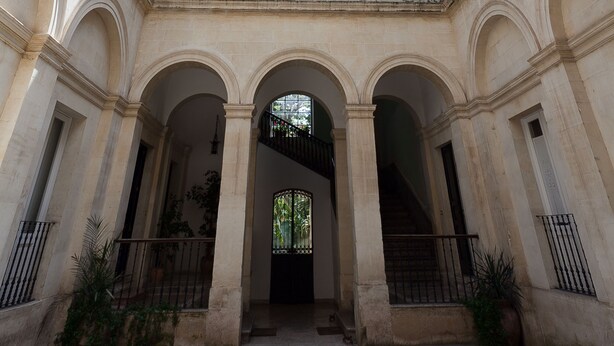 Ein Innenhof mit einem zentralen Bogen, Steinboden und Blick auf ein Gebäude mit zahlreichen Fenstern und Balkonen.