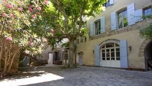 Un patio con pavimento de piedra, un árbol con flores rosadas y un edificio con contraventanas azules y un gran ventanal arqueado.