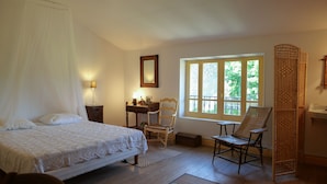 Un dormitorio con cama con dosel, un escritorio con silla, un ventanal con vista a follaje y una silla de mimbre.