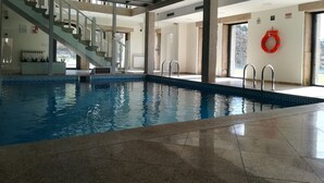Piscina interna com água cristalina, piso ladrilhado e escadaria que leva a um andar superior.