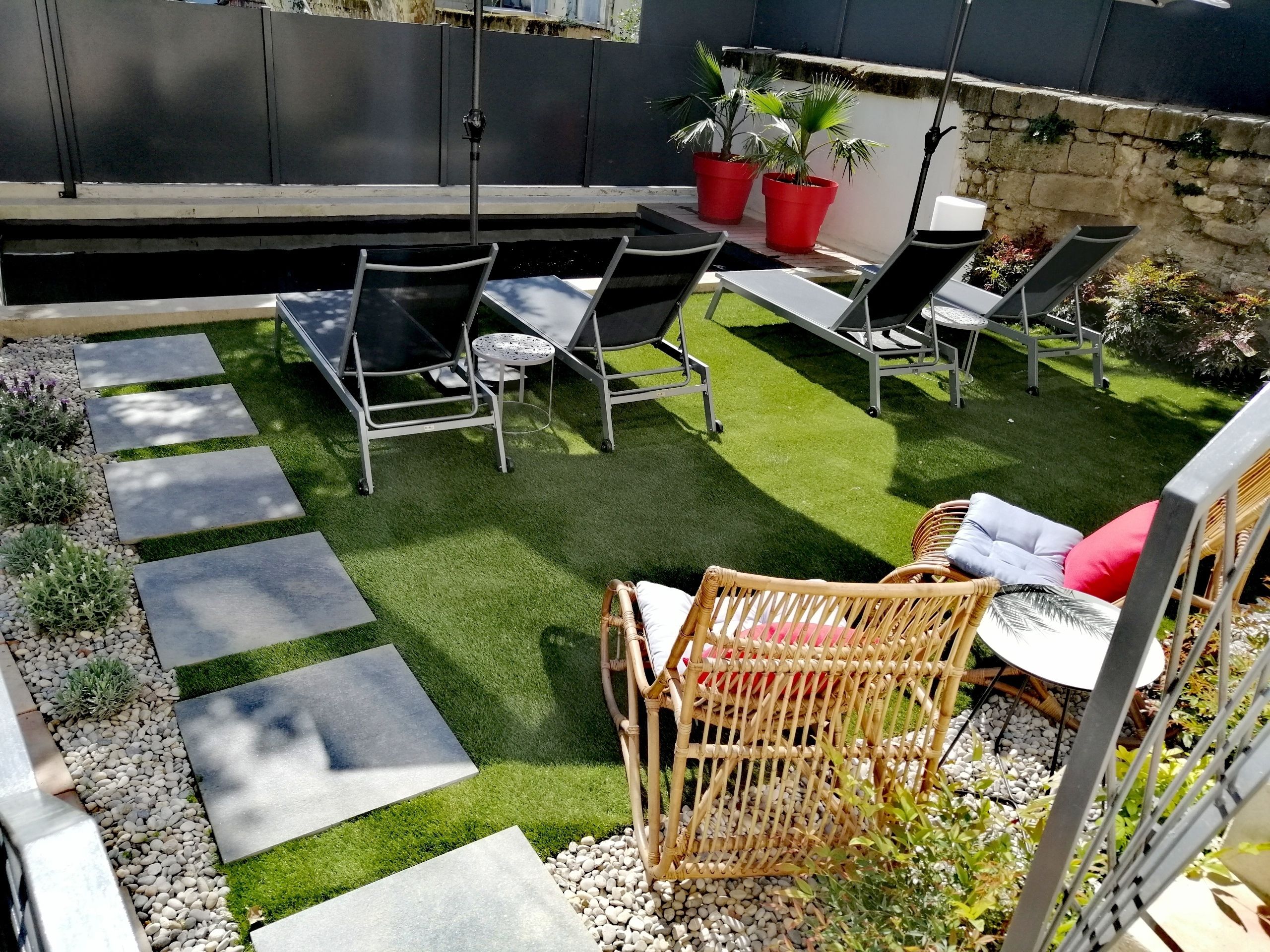 Un patio con mesa y sillas, rodeado de un jardín con césped artificial y senderos de piedra.
