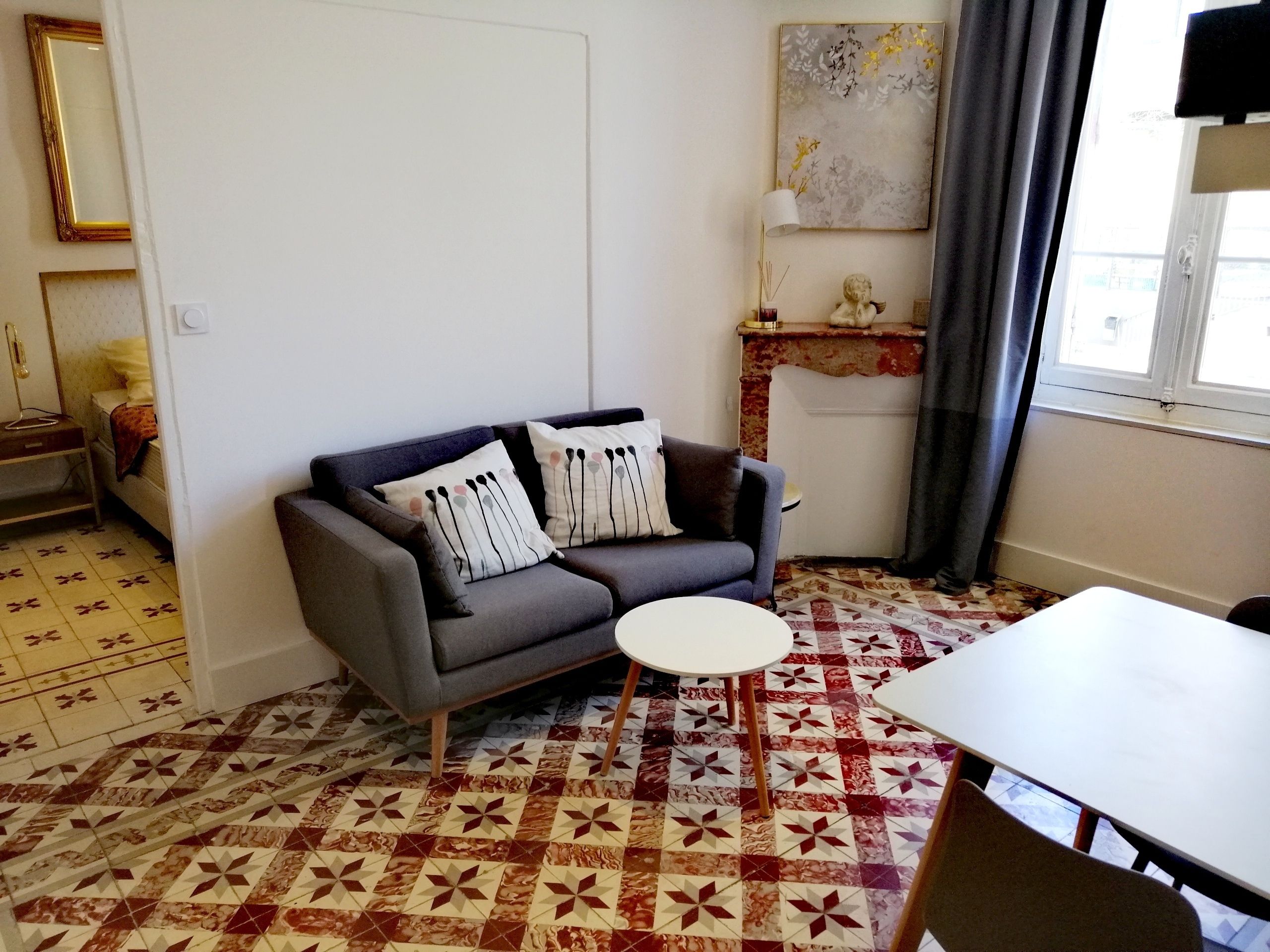 Una sala de estar con un sofá gris, baldosas de suelo con diseño, una mesa blanca y un cuadro en la pared.