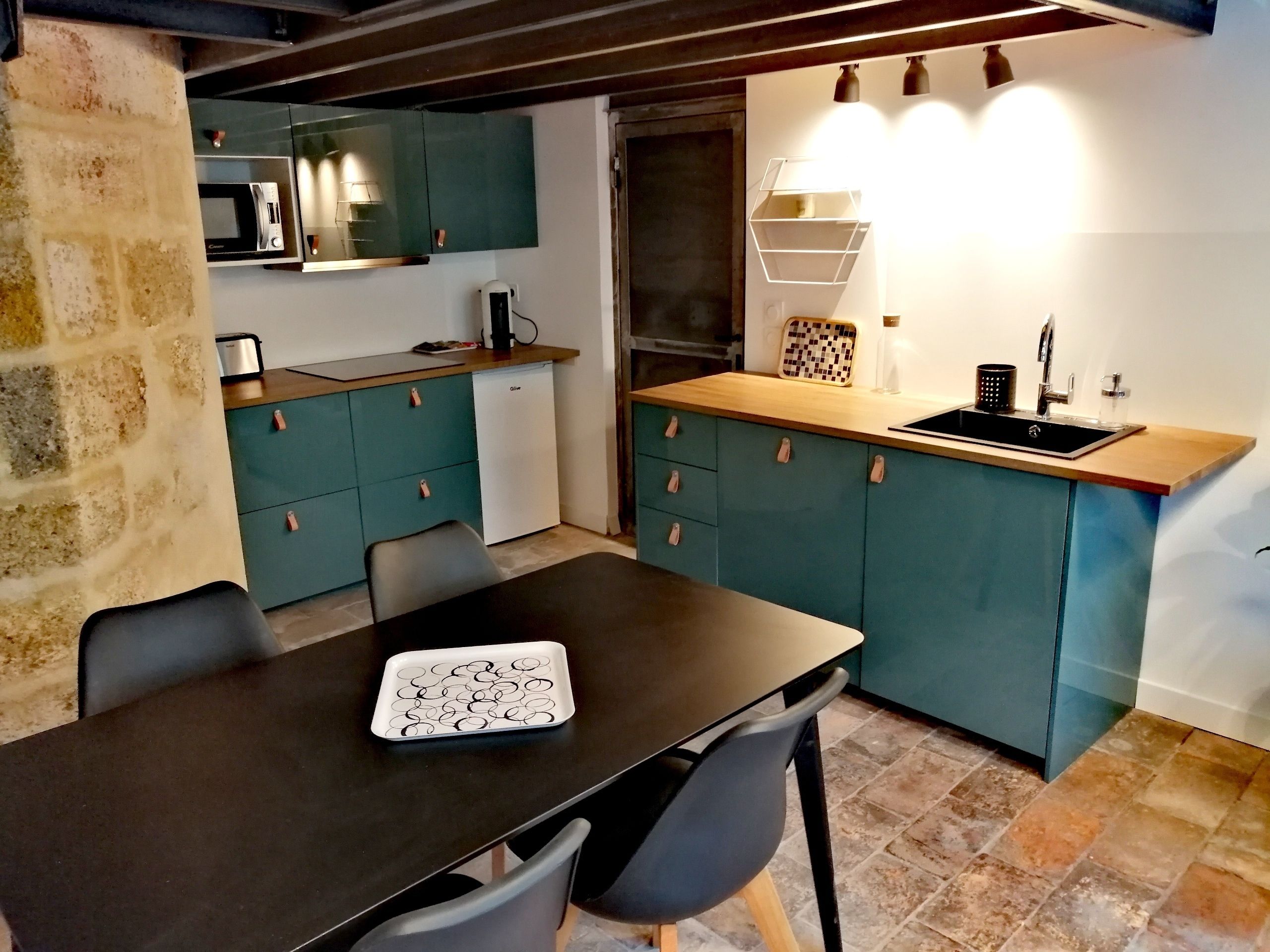 Een kleine keuken met blauwgroene kastjes, een zwart aanrecht en een magnetron. Er is een spoelbak met een kraan, een koffiezetapparaat en een geruit dienblad op het aanrecht.