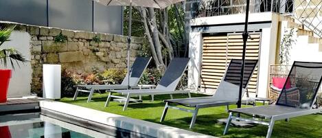 A swimming pool with lounge chairs and umbrellas, surrounded by a stone wall and greenery.
